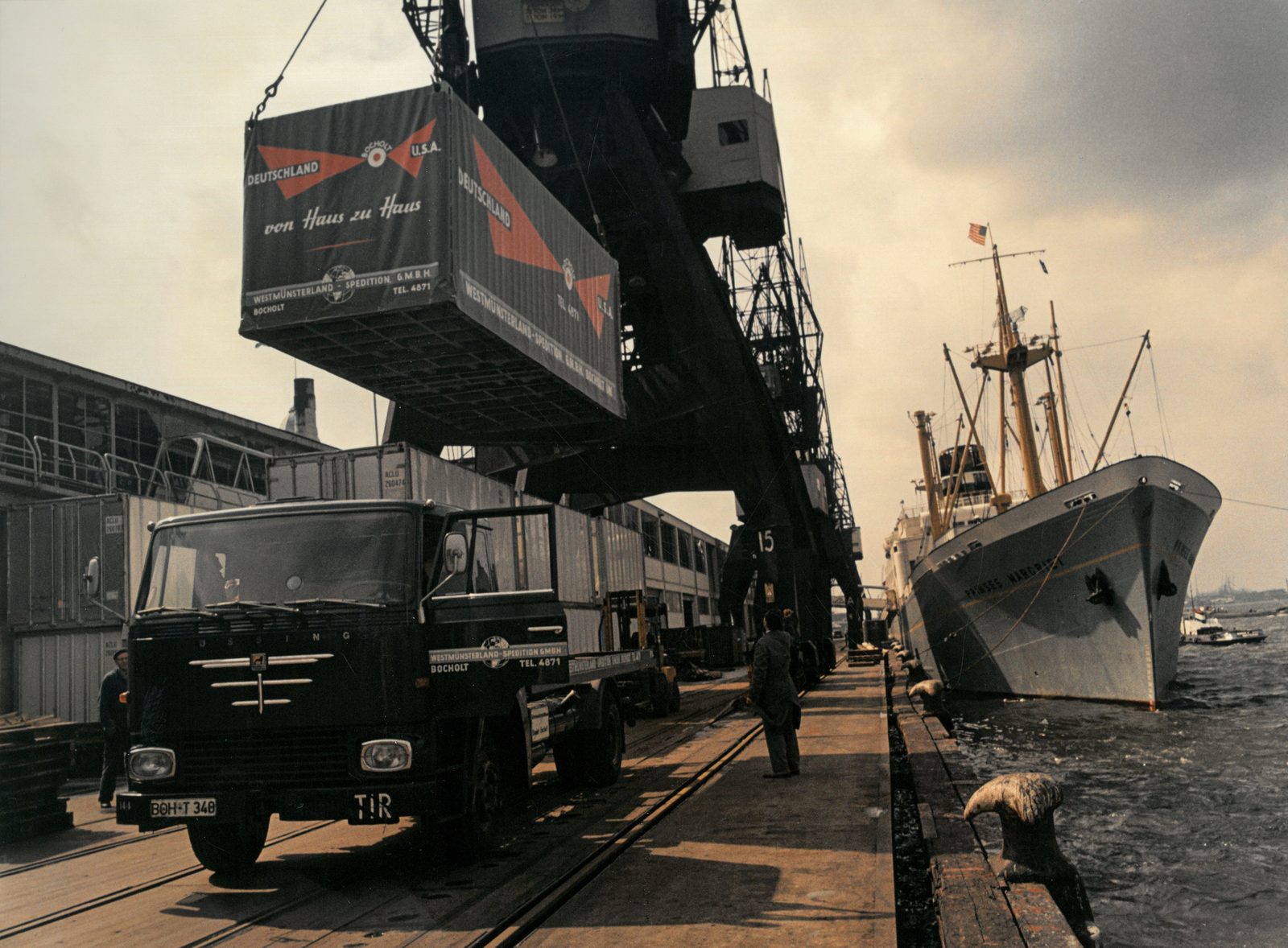 Historische Aufnahme: Der Hafenkran hebt einen Behälter der Westmünsterland-Spedition mit dem Aufdruck „von Haus zu Haus" über einen Lkw, daneben liegt ein Frachtschiff.