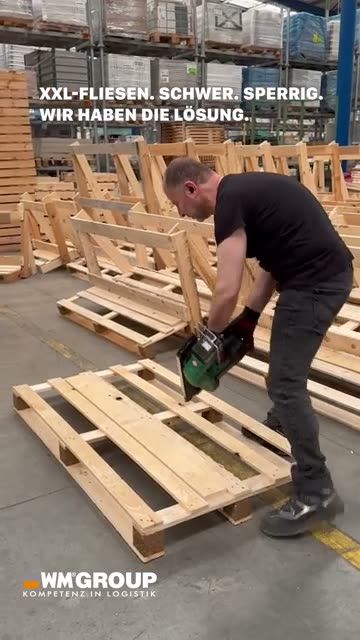 Ein Kollege verschraubt im Lager ein Holzgestell, dahinter stapeln sich fertige Gestelle und Paletten.